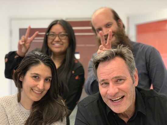 Four people pose indoors; two in the front are smiling at the camera, while the two in the back make peace signs behind the others' heads.