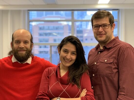 Three people are standing in front of a window with city buildings in the background. They are smiling and wearing red tops. The person on the left has a beard and bald head, the person in the middle has long dark hair, and the person on the right wears glasses.