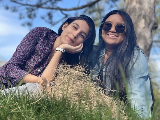 Two women are relaxing on the grass on a sunny day. One is lying on her side, wearing a patterned shirt and smiling with her hand supporting her head. The other is sitting, wearing sunglasses, a denim jacket, and smiling. A tree and a clear sky are in the background.