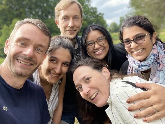 A group of six people, both men and women, are standing outdoors and smiling at the camera. They appear to be friends or colleagues enjoying a sunny day. Trees and greenery are visible in the background.