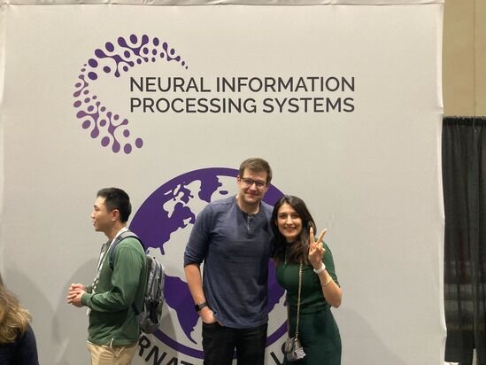 Three people stand in front of a backdrop displaying the 'Neural Information Processing Systems' logo. At the center, two individuals pose for a photo, with the woman on the right making a peace sign. The conference logo is prominently displayed above them.