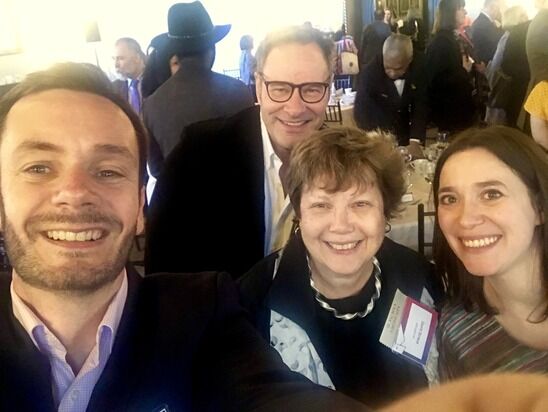 Four people are smiling for a selfie at a social event. They are indoors, with tables and other people in the background. The photo captures a happy and lively atmosphere, with attendees engaged in conversations.
