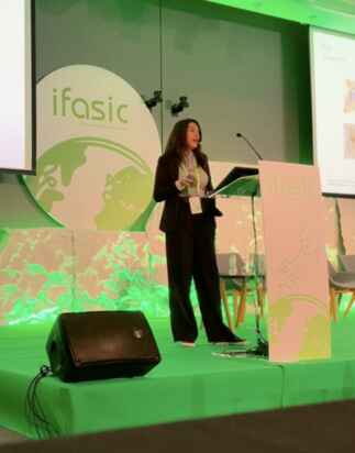 A woman stands on a green-lit stage, delivering a presentation at the ifasic conference. She is speaking into a microphone and is positioned behind a podium with ifasic branding. Two empty chairs and a projection screen are visible in the background.